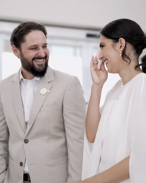 Noivo de terno bege e noiva de branco, os dois rindo; ela enxuga uma lágrima